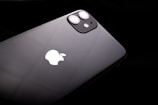 A black smartphone featuring a dual-lens camera and the iconic white apple logo. The device is slightly tilted against a dark background, highlighting its sleek and glossy finish.