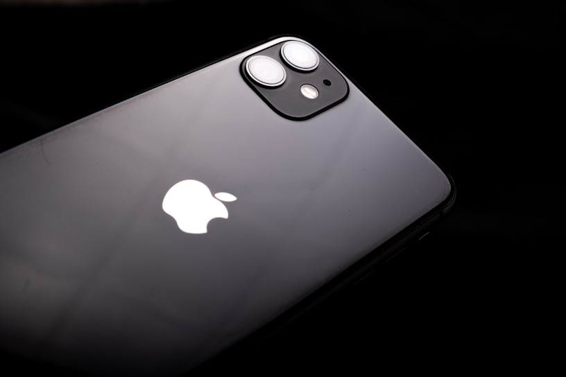 A black smartphone featuring a dual-lens camera and the iconic white apple logo. The device is slightly tilted against a dark background, highlighting its sleek and glossy finish.