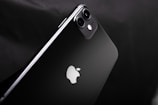 A sleek black smartphone with a dual-camera setup on a glossy surface. The iconic apple logo is prominent in the center. The design is minimalistic and exhibits a modern aesthetic.