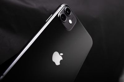 A sleek black smartphone with a dual-camera setup on a glossy surface. The iconic apple logo is prominent in the center. The design is minimalistic and exhibits a modern aesthetic.