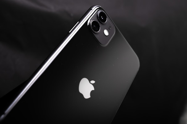 A sleek black smartphone with a dual-camera setup on a glossy surface. The iconic apple logo is prominent in the center. The design is minimalistic and exhibits a modern aesthetic.