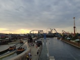 An amusement park features several rides, including a roller coaster and a Ferris wheel. There are multiple sections with water rides and pathways where people are walking. The sky is overcast with a mixture of clouds and a hint of a sunset in the background.
