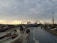 An amusement park features several rides, including a roller coaster and a Ferris wheel. There are multiple sections with water rides and pathways where people are walking. The sky is overcast with a mixture of clouds and a hint of a sunset in the background.