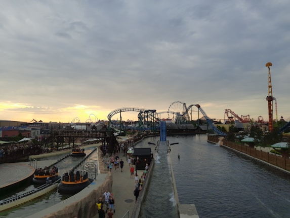 An amusement park features several rides, including a roller coaster and a Ferris wheel. There are multiple sections with water rides and pathways where people are walking. The sky is overcast with a mixture of clouds and a hint of a sunset in the background.