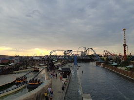 An amusement park features several rides, including a roller coaster and a Ferris wheel. There are multiple sections with water rides and pathways where people are walking. The sky is overcast with a mixture of clouds and a hint of a sunset in the background.