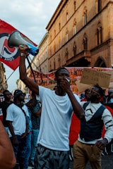 A group of people gathers at a protest, with a person in the foreground holding a megaphone and making a gesture with their hand. Several individuals hold placards with messages, and there is a red flag with a symbol among the crowd. The setting is urban, with a historic building as a backdrop.