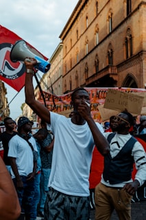 A group of people gathers at a protest, with a person in the foreground holding a megaphone and making a gesture with their hand. Several individuals hold placards with messages, and there is a red flag with a symbol among the crowd. The setting is urban, with a historic building as a backdrop.