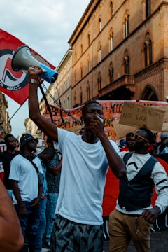 A group of people gathers at a protest, with a person in the foreground holding a megaphone and making a gesture with their hand. Several individuals hold placards with messages, and there is a red flag with a symbol among the crowd. The setting is urban, with a historic building as a backdrop.