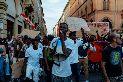 A group of people are marching in a city street, holding signs with slogans like 'Freedom Movement.' The crowd consists of men and women of various ages, some dressed casually, with one individual speaking through a megaphone. The scene is set against a backdrop of historic buildings.