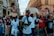 A group of people are marching in a city street, holding signs with slogans like 'Freedom Movement.' The crowd consists of men and women of various ages, some dressed casually, with one individual speaking through a megaphone. The scene is set against a backdrop of historic buildings.