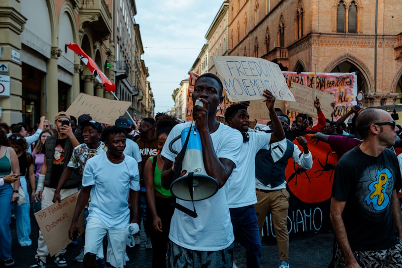 A group of people are marching in a city street, holding signs with slogans like 'Freedom Movement.' The crowd consists of men and women of various ages, some dressed casually, with one individual speaking through a megaphone. The scene is set against a backdrop of historic buildings.