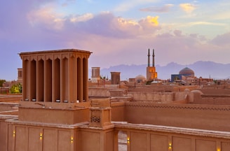An ancient cityscape with traditional windcatchers and adobe buildings. A grand mosque with turquoise domes and tall minarets stands prominently against a backdrop of distant mountains. The sky is painted with a blend of warm sunset hues, adding a serene beauty to the scene.