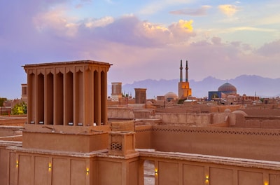 An ancient cityscape with traditional windcatchers and adobe buildings. A grand mosque with turquoise domes and tall minarets stands prominently against a backdrop of distant mountains. The sky is painted with a blend of warm sunset hues, adding a serene beauty to the scene.