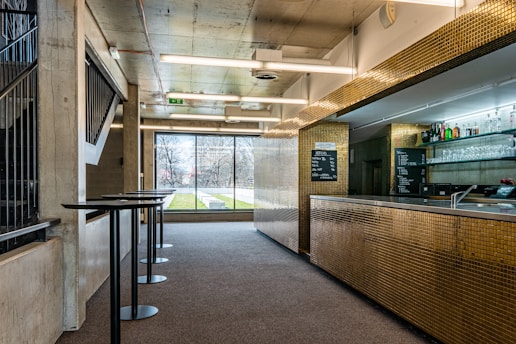 A modern bar area with a sleek metallic counter covered in small golden tiles. Glass shelves behind the counter display various bottles of alcohol and glassware. The space is well-lit with fluorescent lights on the ceiling. Large windows provide a view of an outdoor area with grass and leafless trees. Tall black tables are aligned near the window, and stairs on the left lead to another level.