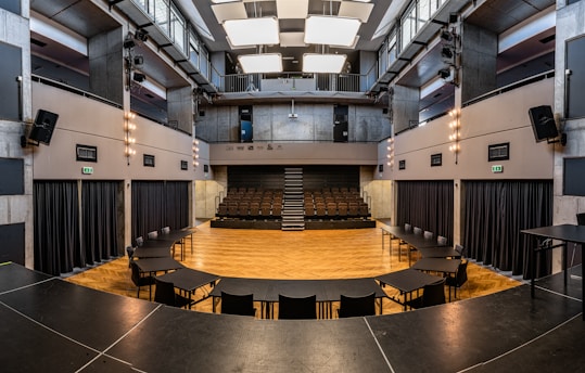 A modern, empty theater or auditorium with a semi-circular arrangement of chairs and tables on a wooden floor. The walls and ceiling are made of gray concrete with exposed lighting and black curtains. Overhead, there are large rectangular light fixtures.