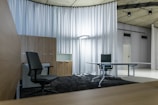 A modern office space with minimalistic design, featuring two office chairs, a sleek table, a storage cabinet, and a floor lamp. The room is decorated with light-colored curtains and has a polished concrete ceiling. The overall ambiance is professional and clean.