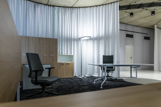 A modern office space with minimalistic design, featuring two office chairs, a sleek table, a storage cabinet, and a floor lamp. The room is decorated with light-colored curtains and has a polished concrete ceiling. The overall ambiance is professional and clean.