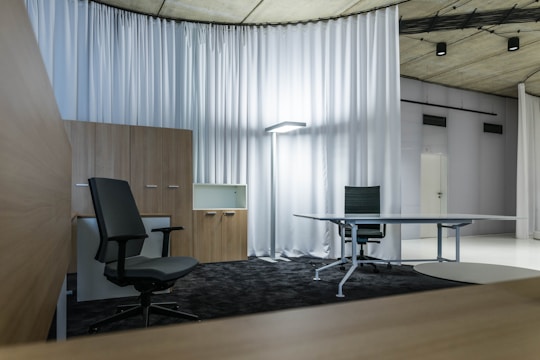 A modern office space with minimalistic design, featuring two office chairs, a sleek table, a storage cabinet, and a floor lamp. The room is decorated with light-colored curtains and has a polished concrete ceiling. The overall ambiance is professional and clean.