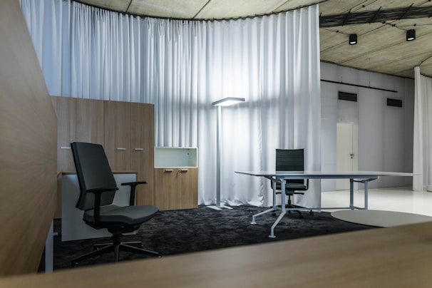 A modern office space with minimalistic design, featuring two office chairs, a sleek table, a storage cabinet, and a floor lamp. The room is decorated with light-colored curtains and has a polished concrete ceiling. The overall ambiance is professional and clean.