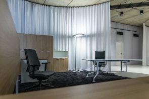 A modern office space with minimalistic design, featuring two office chairs, a sleek table, a storage cabinet, and a floor lamp. The room is decorated with light-colored curtains and has a polished concrete ceiling. The overall ambiance is professional and clean.