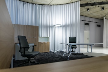 A modern office space with minimalistic design, featuring two office chairs, a sleek table, a storage cabinet, and a floor lamp. The room is decorated with light-colored curtains and has a polished concrete ceiling. The overall ambiance is professional and clean.