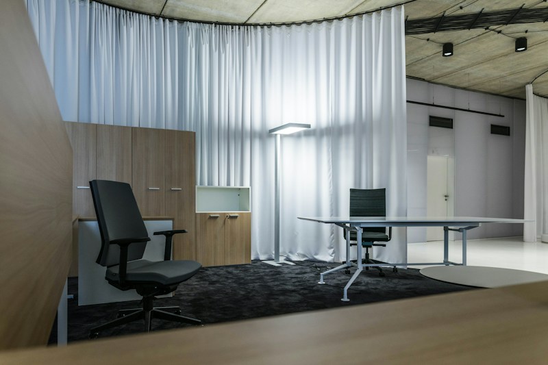 A modern office space with minimalistic design, featuring two office chairs, a sleek table, a storage cabinet, and a floor lamp. The room is decorated with light-colored curtains and has a polished concrete ceiling. The overall ambiance is professional and clean.