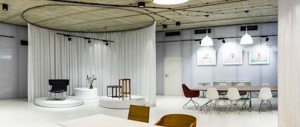 A modern interior space featuring a minimalist design with sleek furniture, including a black chair, a wooden stool, and white circular platforms. The room is well-lit with white and black pendant lights, and the walls have soft white curtains. A long table with white chairs is present, and the decor includes framed artworks on the wall.