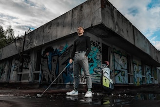 A person stands confidently holding a golf club outside an abandoned building covered in colorful graffiti. They are dressed in a black turtleneck, grey pants, and white sneakers. Next to them is a golf bag with clubs. The scene has a moody sky, with patches of blue showing through the clouds.