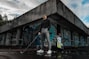 A person stands confidently holding a golf club outside an abandoned building covered in colorful graffiti. They are dressed in a black turtleneck, grey pants, and white sneakers. Next to them is a golf bag with clubs. The scene has a moody sky, with patches of blue showing through the clouds.