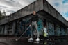 A person stands confidently holding a golf club outside an abandoned building covered in colorful graffiti. They are dressed in a black turtleneck, grey pants, and white sneakers. Next to them is a golf bag with clubs. The scene has a moody sky, with patches of blue showing through the clouds.