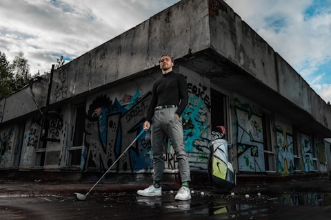 A person stands confidently holding a golf club outside an abandoned building covered in colorful graffiti. They are dressed in a black turtleneck, grey pants, and white sneakers. Next to them is a golf bag with clubs. The scene has a moody sky, with patches of blue showing through the clouds.
