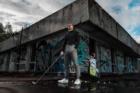 A person stands confidently holding a golf club outside an abandoned building covered in colorful graffiti. They are dressed in a black turtleneck, grey pants, and white sneakers. Next to them is a golf bag with clubs. The scene has a moody sky, with patches of blue showing through the clouds.