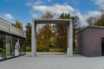 A modern outdoor space with a minimalist concrete structure framing the entrance to a park area. On the left, there is a building with large glass windows displaying books and text in green. The trees around the area suggest an autumn season with foliage in shades of brown and orange.