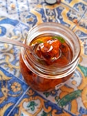 A glass jar filled with a translucent amber-colored preserve, featuring chunks of fruit and a few green leaves. The jar is open, with a metal spoon lifted to reveal a portion of the preserve. It rests on a colorful, intricately patterned tablecloth dominated by blue, yellow, and orange colors.