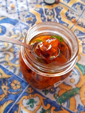 A glass jar filled with a translucent amber-colored preserve, featuring chunks of fruit and a few green leaves. The jar is open, with a metal spoon lifted to reveal a portion of the preserve. It rests on a colorful, intricately patterned tablecloth dominated by blue, yellow, and orange colors.