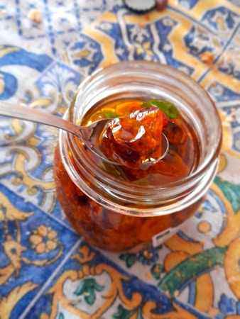 A glass jar filled with a translucent amber-colored preserve, featuring chunks of fruit and a few green leaves. The jar is open, with a metal spoon lifted to reveal a portion of the preserve. It rests on a colorful, intricately patterned tablecloth dominated by blue, yellow, and orange colors.