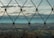 A wire mesh fence is in clear focus with a blurred landscape of rolling hills and a cloudy sky in the background.