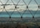 A wire mesh fence is in clear focus with a blurred landscape of rolling hills and a cloudy sky in the background.