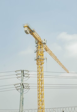 A tall yellow construction crane stretches into the sky, situated near power lines and framed against a partly cloudy blue sky. The crane stands upright, towering over the electrical poles visible below.