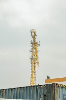 A tall yellow construction crane stands prominently against a cloudy sky. The base of the crane is positioned behind industrial metal structures or containers. The crane's metal structure features multiple platforms and ladders, exemplifying typical construction site elements.