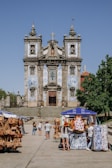 A historic church adorned with intricate blue and white azulejos stands as the focal point. In the foreground, a market scene unfolds with people browsing stalls that display colorful textiles and leather goods. The sky is clear, and a tree provides a touch of greenery to the right.