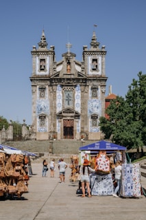 A historic church adorned with intricate blue and white azulejos stands as the focal point. In the foreground, a market scene unfolds with people browsing stalls that display colorful textiles and leather goods. The sky is clear, and a tree provides a touch of greenery to the right.