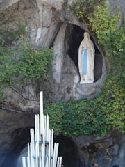 A religious statue of the Virgin Mary is set within a rocky grotto, surrounded by lush green vines and foliage. In front of the statue, a large arrangement of tall, white candles is visible, adding a solemn and sacred atmosphere.