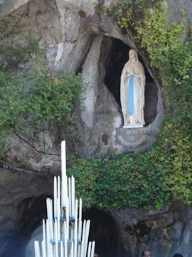 A religious statue of the Virgin Mary is set within a rocky grotto, surrounded by lush green vines and foliage. In front of the statue, a large arrangement of tall, white candles is visible, adding a solemn and sacred atmosphere.