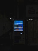 A vending machine is positioned in a dimly lit urban setting, surrounded by shadows. The machine is brightly illuminated, highlighting its colorful array of beverages behind the glass display. The contrast between the light of the machine and the surrounding darkness is stark.