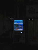 A vending machine is positioned in a dimly lit urban setting, surrounded by shadows. The machine is brightly illuminated, highlighting its colorful array of beverages behind the glass display. The contrast between the light of the machine and the surrounding darkness is stark.