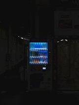 A vending machine is positioned in a dimly lit urban setting, surrounded by shadows. The machine is brightly illuminated, highlighting its colorful array of beverages behind the glass display. The contrast between the light of the machine and the surrounding darkness is stark.