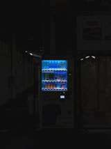 A vending machine is positioned in a dimly lit urban setting, surrounded by shadows. The machine is brightly illuminated, highlighting its colorful array of beverages behind the glass display. The contrast between the light of the machine and the surrounding darkness is stark.