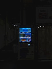 A vending machine is positioned in a dimly lit urban setting, surrounded by shadows. The machine is brightly illuminated, highlighting its colorful array of beverages behind the glass display. The contrast between the light of the machine and the surrounding darkness is stark.