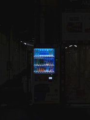 A vending machine is positioned in a dimly lit urban setting, surrounded by shadows. The machine is brightly illuminated, highlighting its colorful array of beverages behind the glass display. The contrast between the light of the machine and the surrounding darkness is stark.